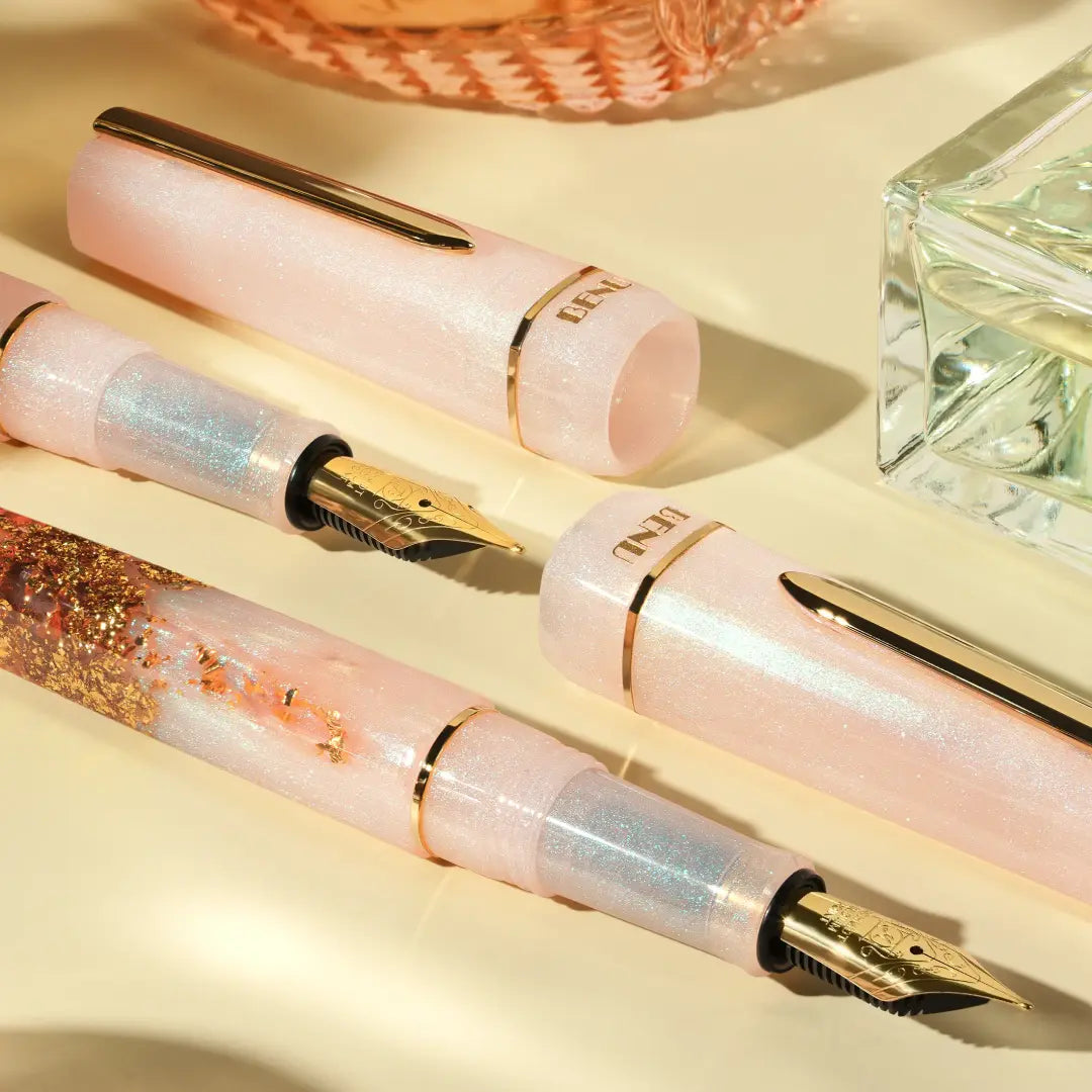 BENU presents the newest Haute Collection — a line of elegant, fashion-inspired pens that blend classic charm with bold, modern flair. The designs include both subtle and vibrant styles, showcasing BENU’s signature glitter and color artistry. Each pen features a faceted body with alternating wide and slender facets, complemented by either gold-plated or chrome-plated trims for a sleek, timeless look.
A mix of golden flakes dances through a soft, pearly pink base, complemented by gleaming gold trim. Tender