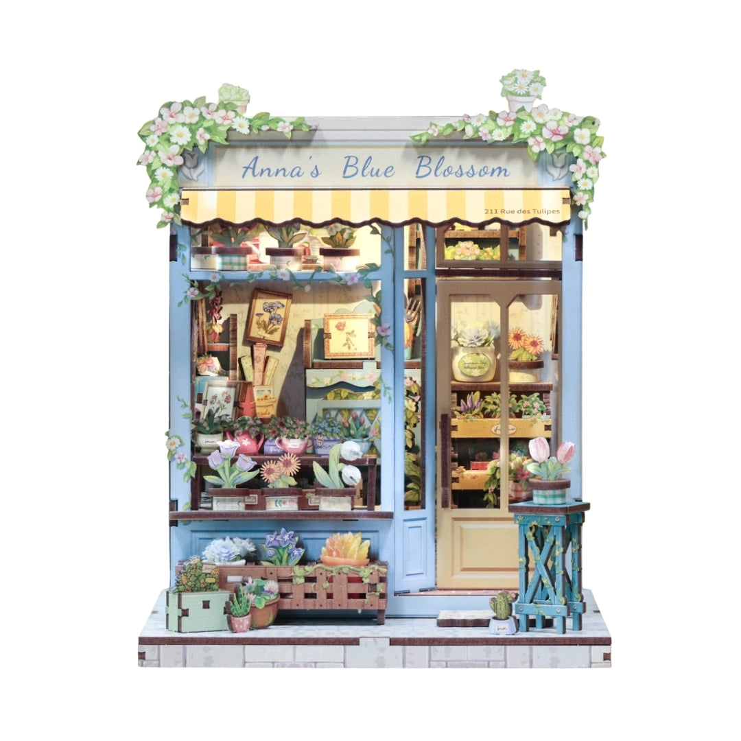 Step into the charm of a quaint flower shop with the Anna's Blue Blossom DIY Miniature Book Nook Kit. From the soft pastel storefront to the cheerful yellow door and overflowing baskets of vibrant blooms, every detail captures the essence of a dreamy neighborhood florist. Inside, shelves brim with blossoms, potted plants, and tiny gardening treasures, creating a warm and welcoming scene. The delicate floral canopy and cascading greenery add a fresh touch of life, making this kit feel like a stroll through a
