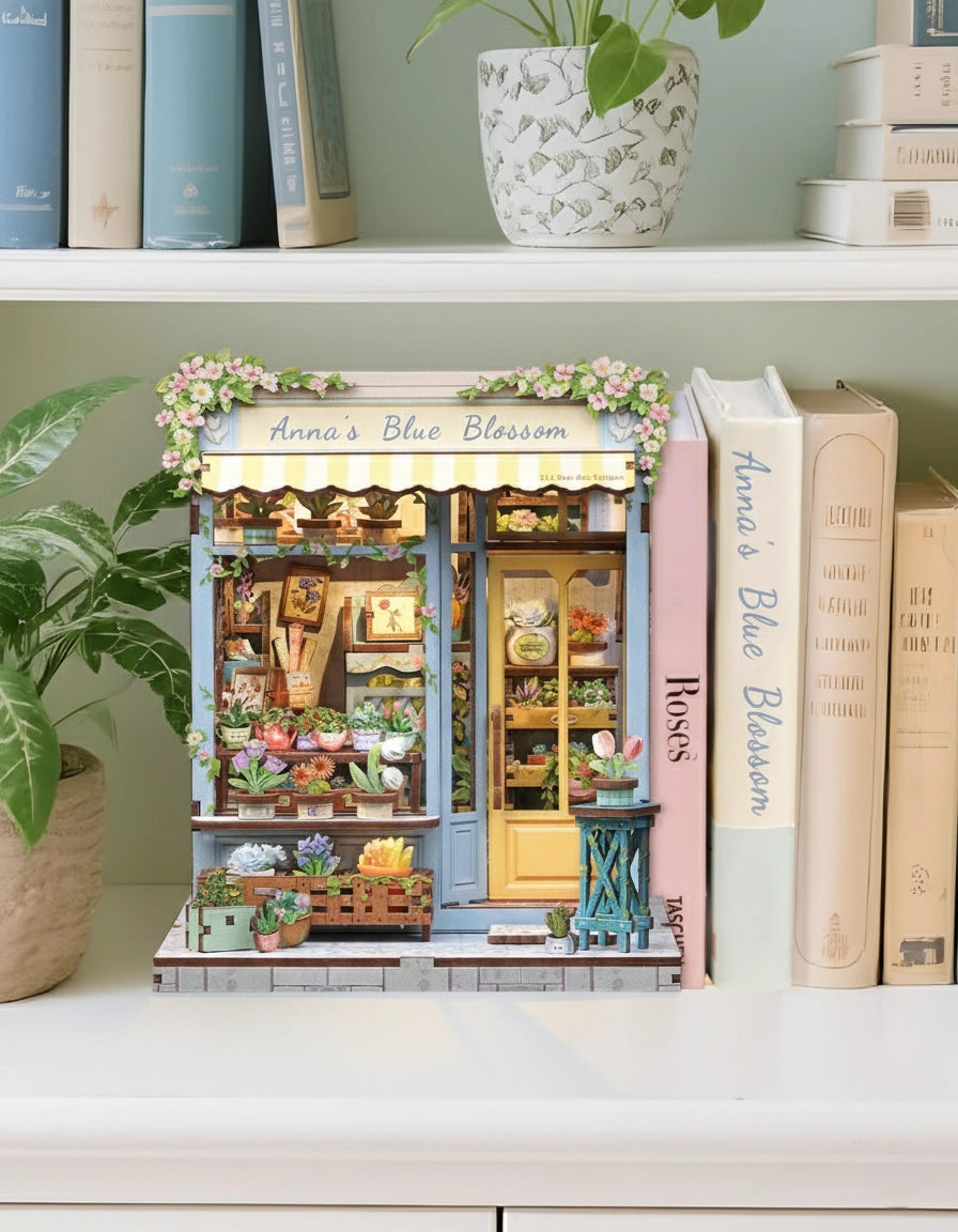 Step into the charm of a quaint flower shop with the Anna's Blue Blossom DIY Miniature Book Nook Kit. From the soft pastel storefront to the cheerful yellow door and overflowing baskets of vibrant blooms, every detail captures the essence of a dreamy neighborhood florist. Inside, shelves brim with blossoms, potted plants, and tiny gardening treasures, creating a warm and welcoming scene. The delicate floral canopy and cascading greenery add a fresh touch of life, making this kit feel like a stroll through a
