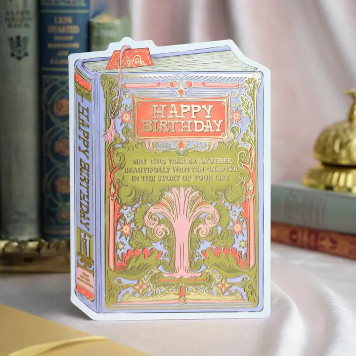 A bookish birthday card for lovers of stories and symbolism. This periwinkle card features a lush, gold-foiled tree with an open book at its roots and the message “May this year be another beautifully written chapter in the story of your life.” A thoughtful and elegant foil-pressed stationery piece.
Product Details:
- Dimensions: 5" x 7"
- Die-cut antique book-style design
- Hand-illustrated with gold foil details
- Blank interior
- Offset and foil printed
- Comes with envelope