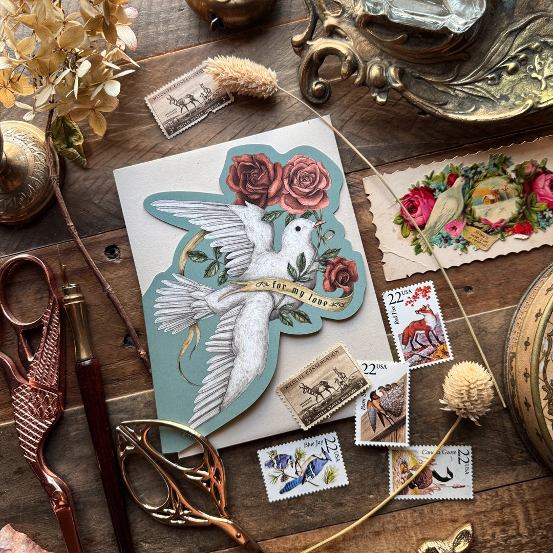 Folding card featuring a dove and roses, illustrated by Jessica Roux. Both doves and roses are symbols of romantic love, and this card is perfect for Valentine's Day, an anniversary, or to tell someone you love them. This card is inspired by Victorian Valentine cards.
Single card with envelope.
A2 size card in a cut-out shape of a dove with roses
Folded
Blank on the inside
Comes with a kraft paper envelope. Packaged in a cello sleeve.
Luxurious, soft-touch coating