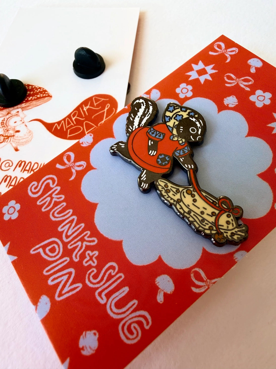 A Skunk and their friend the Slug take a walk through the forest.
This hard enamel pin measures approximately 1.75" and features a rubber clasp for easy closure and two back posts so pin stays where you stick it. Comes with a back card.