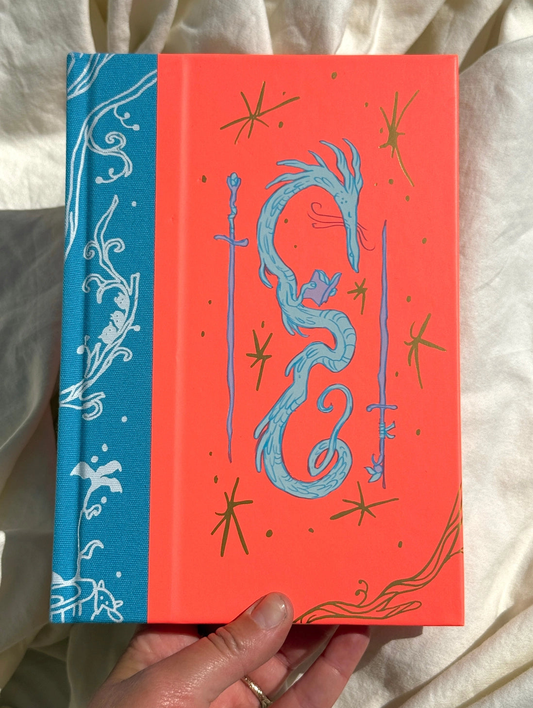 This is a blank 200 page notebook/sketchbook with thin ivory Munken paper perfect for light sketching or writing. Part of a fantasy collection called Woodfolk.
The cover is fluorescent pink/orange and has a smooth matte finish with gold foiling on the front and back. Plus it has a blue fabric spine with white embossing, adding to the charm of the book. Best for pencils, light pens, and generally light mediums, as thick markers or water can bleed. Not recommended for gouache or heavy marker use.