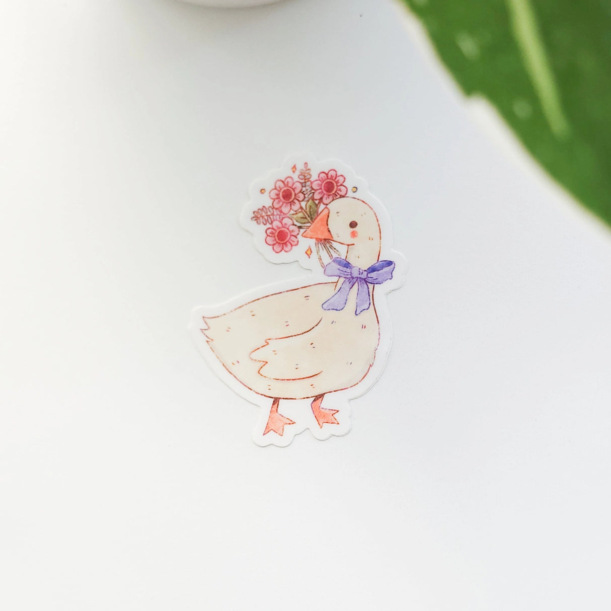 A sweet goose pauses with a bouquet of flowers held gently in its beak, a blue bow tied neatly at its neck. This vinyl sticker captures that charming, flower-filled moment, adding a warm, cottage-inspired touch to your everyday essentials.
Made from durable, waterproof vinyl, this sticker is designed to hold up wherever you place it—whether that’s a water bottle, laptop, or journal you reach for daily.