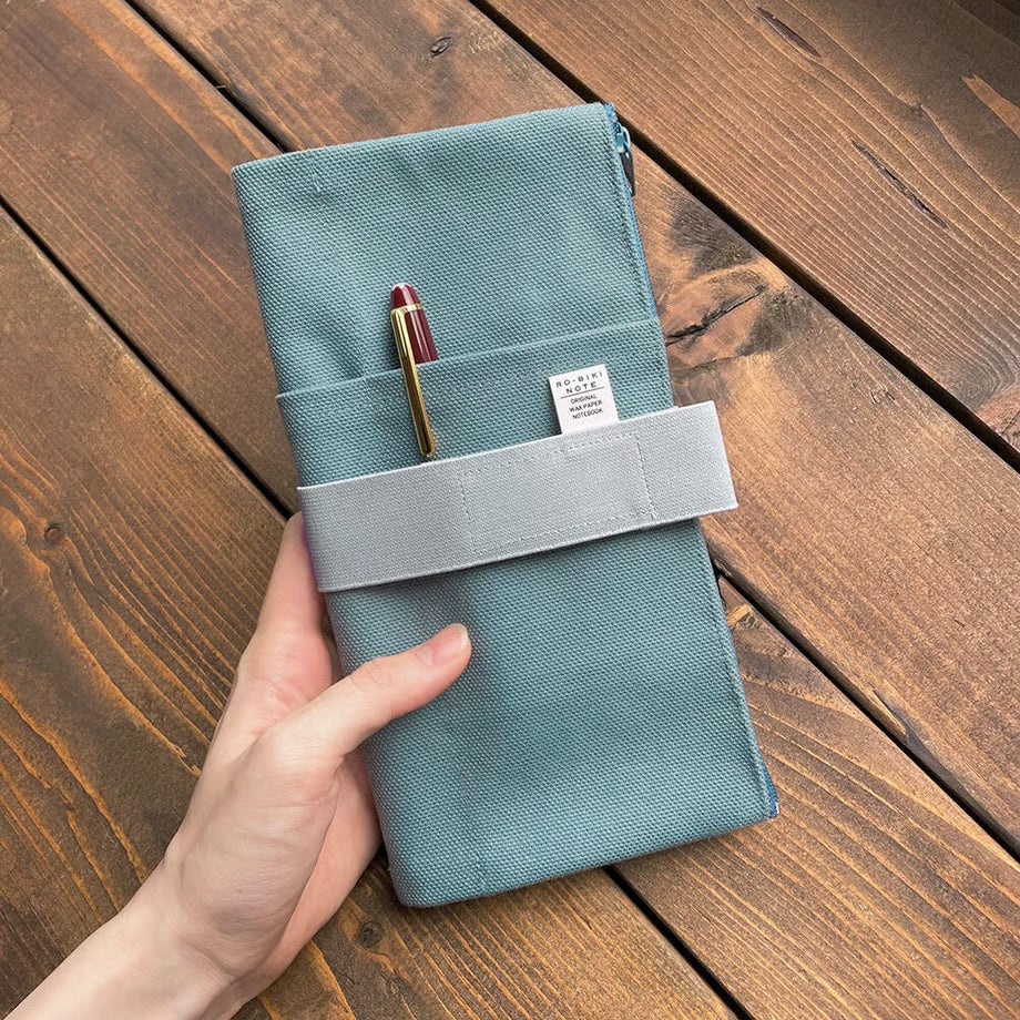 Made of cotton that feels good to the touch, this is RO-BIKI NOTE CANVAS COVER vintage blue. A zippered pocket on the side is useful for storing stationery that tends to be loose. It has a simple design that does not stand out when carrying. When closed, it feels neat and sizable. The surface of the cover has three pockets of different sizes, so you can enjoy multiple storage options.
With a connecting band, you can sandwich the Basic size and the passport size at the same time.