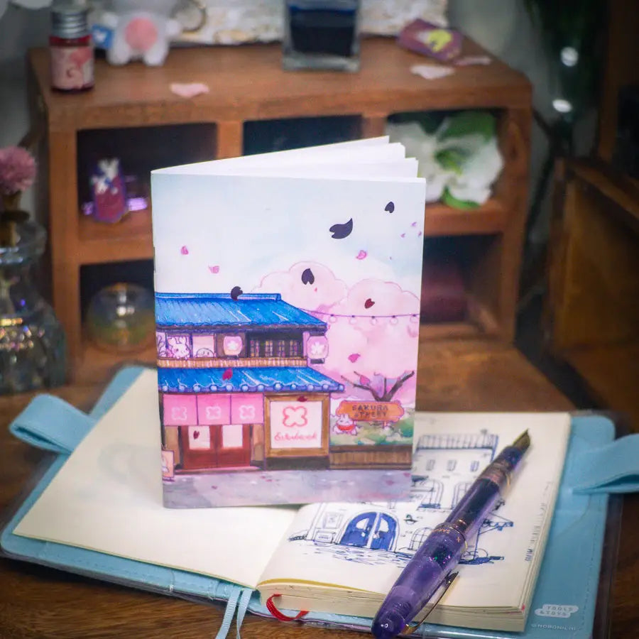 Sakuradragon - Sakura Street Shopfronts A6 Notebook
Cozy little shop fronts that you will find on your walk down Sakura Street. Did you notice the pink foil cherry blossoms falling from the sky?
Dimensions: A6 (10.5 × 14.8 cm)
Paper: Double-sided offset paper
Page Ruling: Blank
Pages: 64
Message from sakuradragon: Not all paper and ink are made equal but this paper does a great job with both fountain pens and dip pens. There is minimal ghosting but bleeding does occur with heavy ink usage.