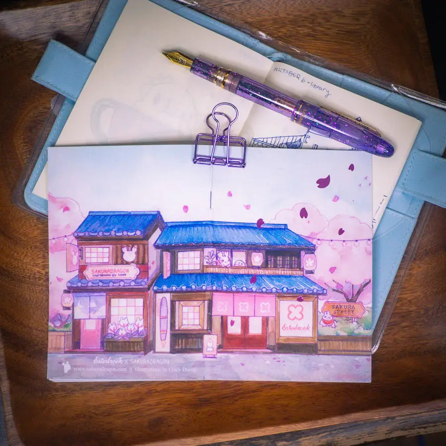 Sakuradragon - Sakura Street Shopfronts A6 Notebook
Cozy little shop fronts that you will find on your walk down Sakura Street. Did you notice the pink foil cherry blossoms falling from the sky?
Dimensions: A6 (10.5 × 14.8 cm)
Paper: Double-sided offset paper
Page Ruling: Blank
Pages: 64
Message from sakuradragon: Not all paper and ink are made equal but this paper does a great job with both fountain pens and dip pens. There is minimal ghosting but bleeding does occur with heavy ink usage.