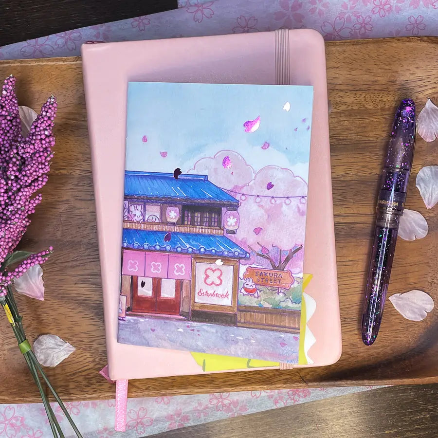 Sakuradragon - Sakura Street Shopfronts A6 Notebook
Cozy little shop fronts that you will find on your walk down Sakura Street. Did you notice the pink foil cherry blossoms falling from the sky?
Dimensions: A6 (10.5 × 14.8 cm)
Paper: Double-sided offset paper
Page Ruling: Blank
Pages: 64
Message from sakuradragon: Not all paper and ink are made equal but this paper does a great job with both fountain pens and dip pens. There is minimal ghosting but bleeding does occur with heavy ink usage.