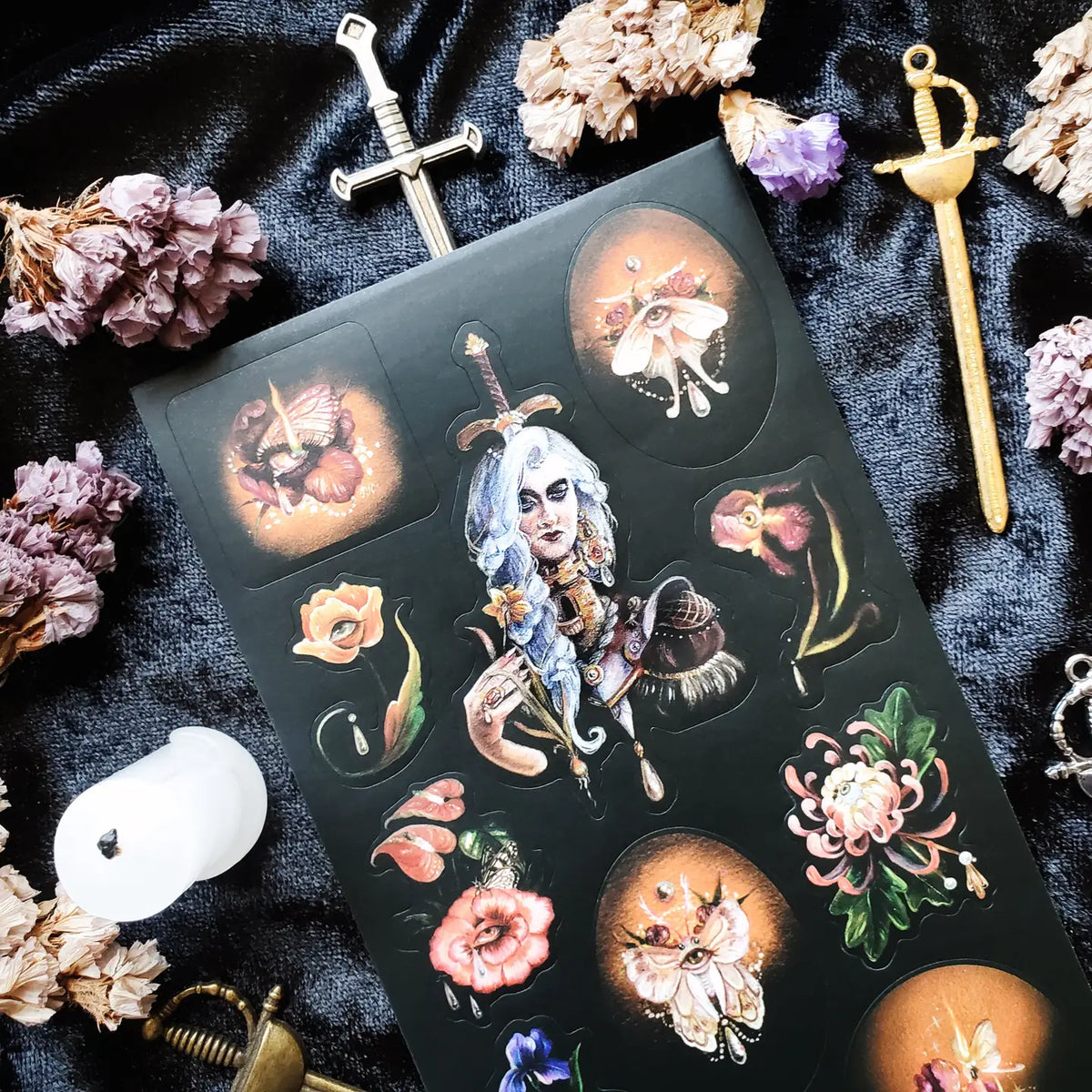 In @iratxe.yc’s shadowed garden, nothing is quite as it first appears. Jeweled flowers gaze back from curling stems, delicate moths wear watchful eyes, and a silver-haired guardian stands adorned in blossoms, gemstones, and armor. Rich with surreal beauty and dark botanical magic, this striking sheet reveals a forest where every petal may be hiding a secret.
Art by: @iratxe.yc
One sticker sheet is included.
Top Coating: None
Adhesive Layer: Paper
Backing Layer: Clear PET
Size:6.69"x3.54"
Weight-0.03oz