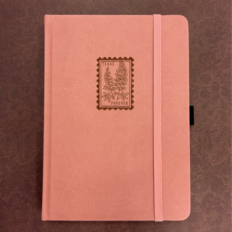 Archer and Olive A5 Notebook - Texas Forever

A charming pink journal featuring a Texas bluebonnet forever stamp on the cover — a sweet nod to the Lone Star State and a perfect keepsake for writers, travelers, and stationery lovers.

✨ Features You’ll Love

Ultra-thick paper designed to prevent ghosting and bleeding

Elastic closure to keep pages secure

Built-in pen loop for your favorite pen or pencil

📓 Notebook Details

Size: A5&nbsp;

Pages:&nbsp;118 pages

Paper:&nbsp;160gsm ultra-thick, bleed-resist