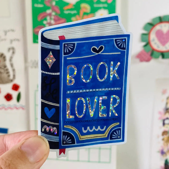 The "Book Lover" glitter sticker from Daria Solak Illustrations is a must-have for all bibliophiles. With a size of 9 cm, it's perfect for adorning your favorite notebook, laptop, or anywhere else you want to express your love for books. Drawing inspiration from everyday life, this sticker is created with hand-drawn illustrations, reflecting Daria Solak's passion for color and design. Add a touch of whimsy and personal style to your items with this vibrant sticker.