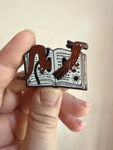 These pins are a lovely, small and cute piece to show off on your clothes, book bags, or wherever you show off your pins!

1.75 inches across, black metal lapel pin with "hard" enamel colors. Black rubber clasps. Packaged in a small plastic bag with a card backing.

"Are your customers book hoarders? No one loves a good horde like a dragon. And I love a good pun, so I combined the two into this Book Wyrm design! Wyrm is the old English word for dragon."