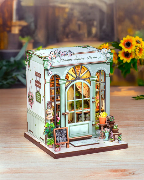 This elegant miniature captures the beauty of a boutique flower shop, adorned with delicate blooms, pastel accents, and a graceful arched doorway that invites you inside.

From the dainty floral arrangements to the hand-painted shop signs and warm, glowing interior, every detail evokes the feeling of strolling down the Champs-Élysées on a sunny afternoon. The soft LED lights cast a welcoming glow, highlighting rows of flowers ready to be wrapped for someone special.
