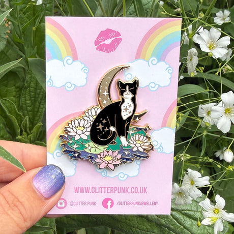 This super sweet fantasy pin features a black and white tuxedo cat relaxing on a lily pad in front of a crescent moon.

It’s 45mm (1.77”) wide, and is made from gunmetal hard enamel, with glitter and UV printed details. It's secured with 2 pink clutches to keep the pin in place.