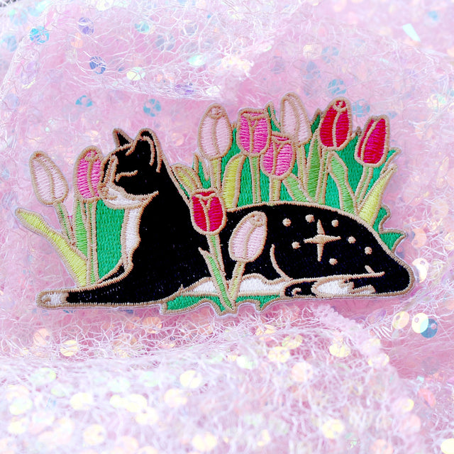 Cats love nothing better than relaxing in a flower bed – and this cute cat is no exception! This awesome embroidered patch features a black and white tuxedo cat lying down in some pink tulips.

Beautifully woven with black, white, pink, green and gold threads, this fabulous patch with brighten up any coat, bag or hat!

Measurements: 100mm wide