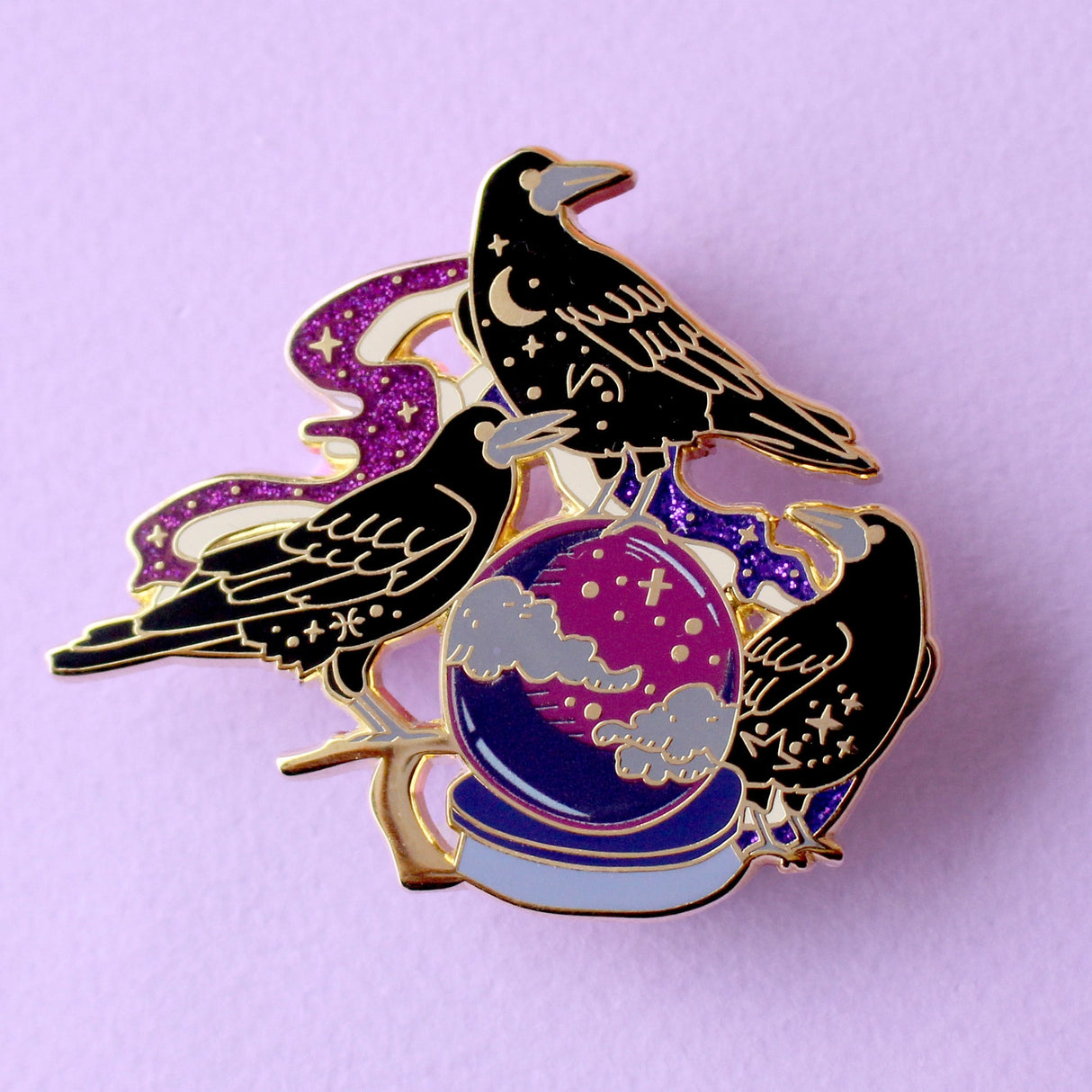 This enamel pin features three rooks sitting around a crystal ball, surrounded by glittery mystical smoke trails. The pin is presented on a Glitter Punk backing card and is secured with 2 pin backs.

This pin is part of the Corvid Collection, an 8-part series of bird pins. 

Measurements: 54mm