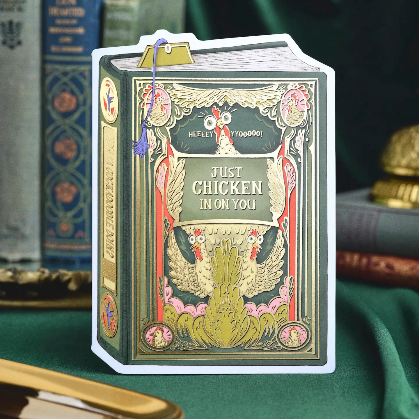 A humorous book-style greeting card that’s sure to spark a smile. This dark green and yellow vintage design features hand-illustrated chickens framed in ornate gold detailing with the playful phrase “Just chickin’ in on you.” A witty and collectible foil-pressed stationery piece that adds a touch of humor to your display.

Product Details:
- Dimensions: 5" x 7"
- Die-cut vintage book cover
- Hand-illustrated with gold foil details
- Blank interior
- Offset and foil printed
- Envelope included