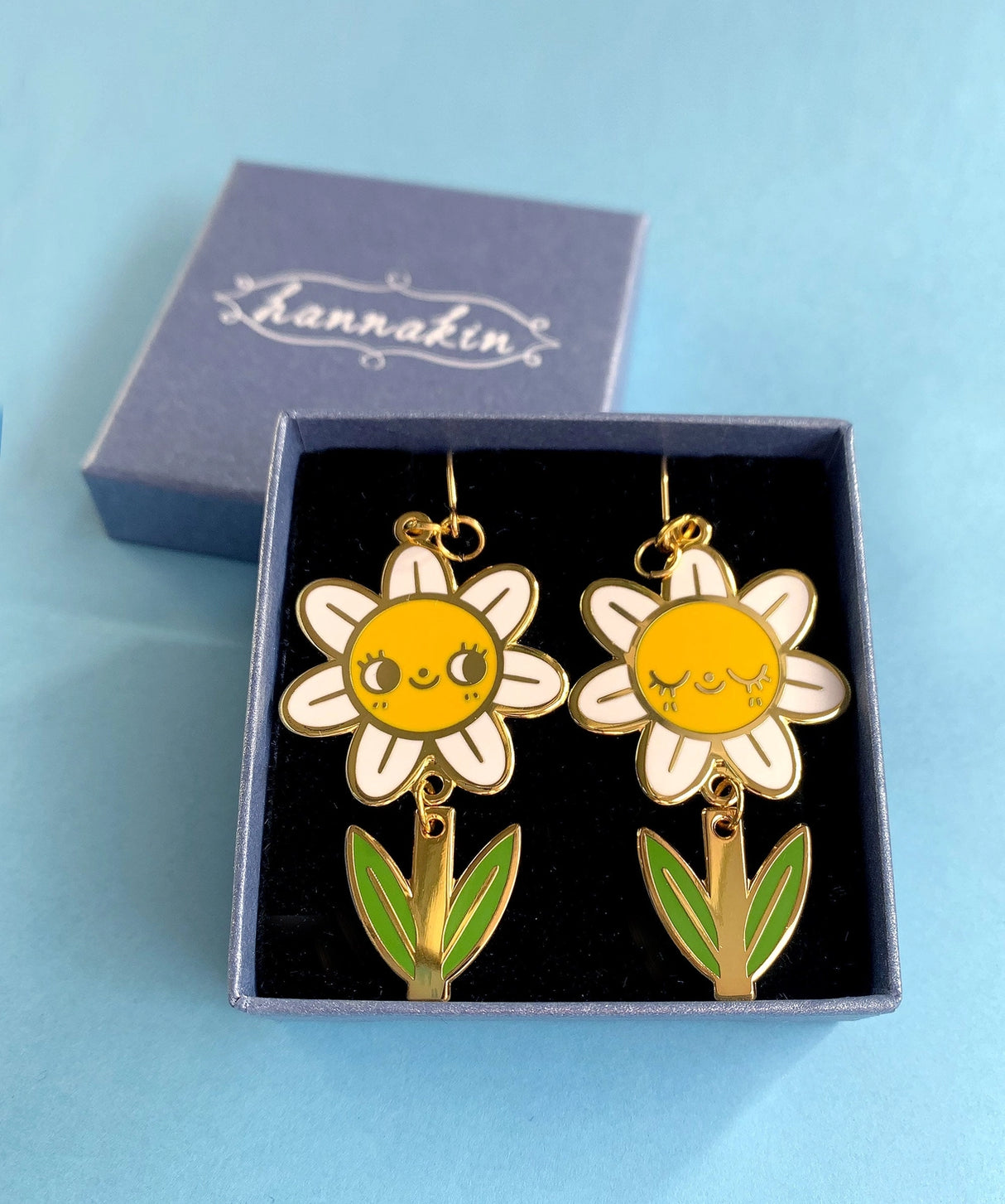A pair of mis-matched happy daisy themed earrings (one one with happy open eyes, and one with happy closed) with separate dangling stems. Made from hard enamel with shiny gold metal finish on high quality Hypo-Allergenic, gold plated, brass based hooks.

Each earring measures approximately 5cm tall x 3cm wide. Earrings come in a Hannakin embossed gift box.