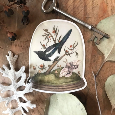Vinyl semi-transparent sticker of One for Sorrow, an illustration of a magpie and moth under a cloche jar, by Jessica Roux.

The glass of the jar is transparent, but the contents and base of the jar are opaque. Sticker measures 2.6" x 3.1" and is glossy.