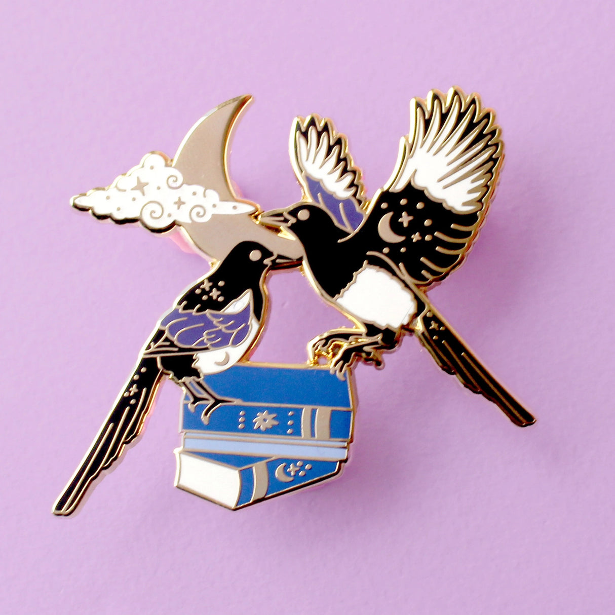 This enamel pin features a pair of magpies (2 for luck!) on top of a set of blue books, in front of a moon and glittery cloud. The pin is presented on a Glitter Punk backing card and is secured with 2 pin backs.

This pin is part of the Corvid Collection, an 8-part series of bird pins. 

Measurements: 55mm