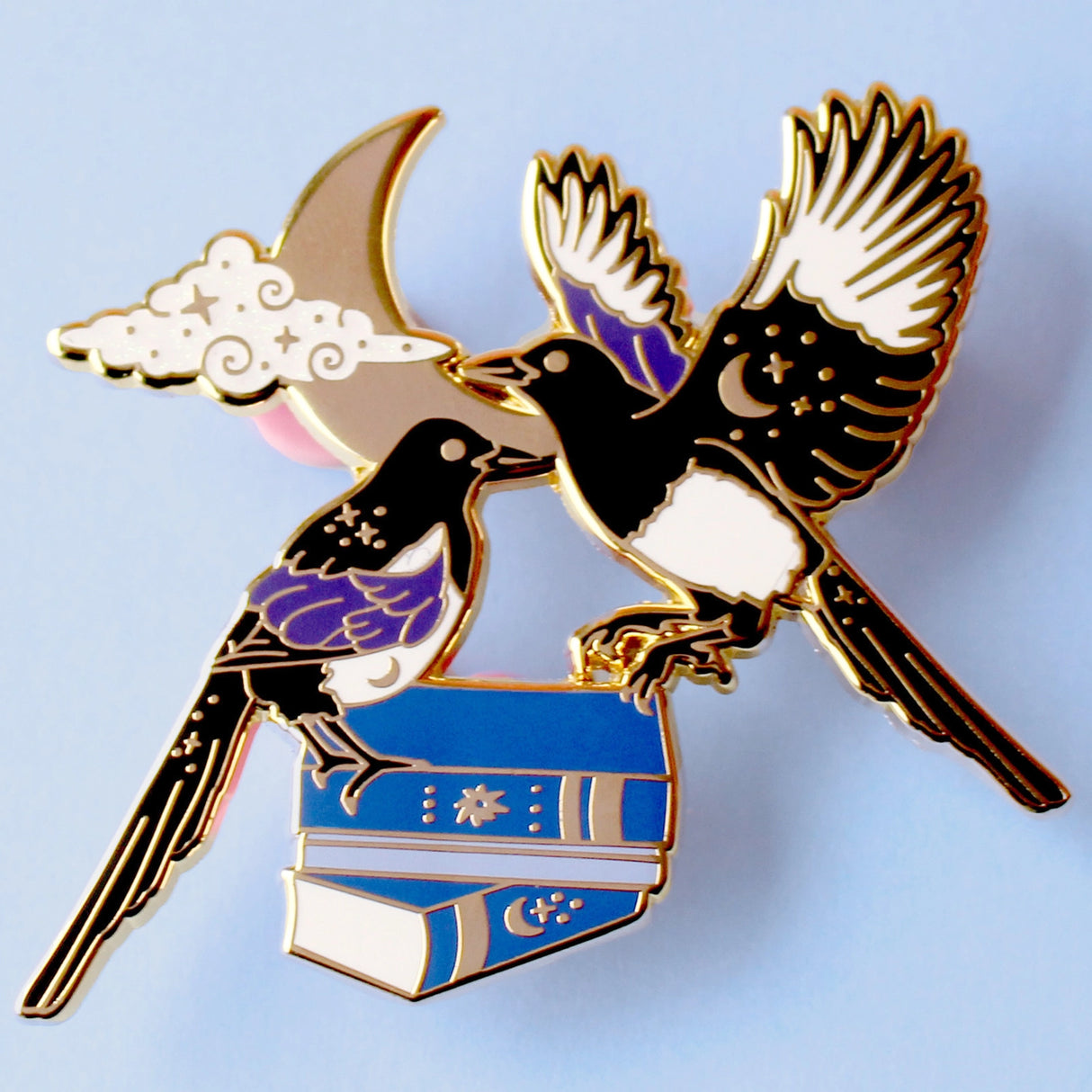 This enamel pin features a pair of magpies (2 for luck!) on top of a set of blue books, in front of a moon and glittery cloud. The pin is presented on a Glitter Punk backing card and is secured with 2 pin backs.

This pin is part of the Corvid Collection, an 8-part series of bird pins. 

Measurements: 55mm