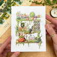 Postcard featuring my original watercolor and ink illustration of the various plants, creatures, and beautiful books nestled on the shelf of a garden witch, including a tiny frog and hedgehog enjoying some itty bitty books, stained glass greenhouses, and endless climbing vines.