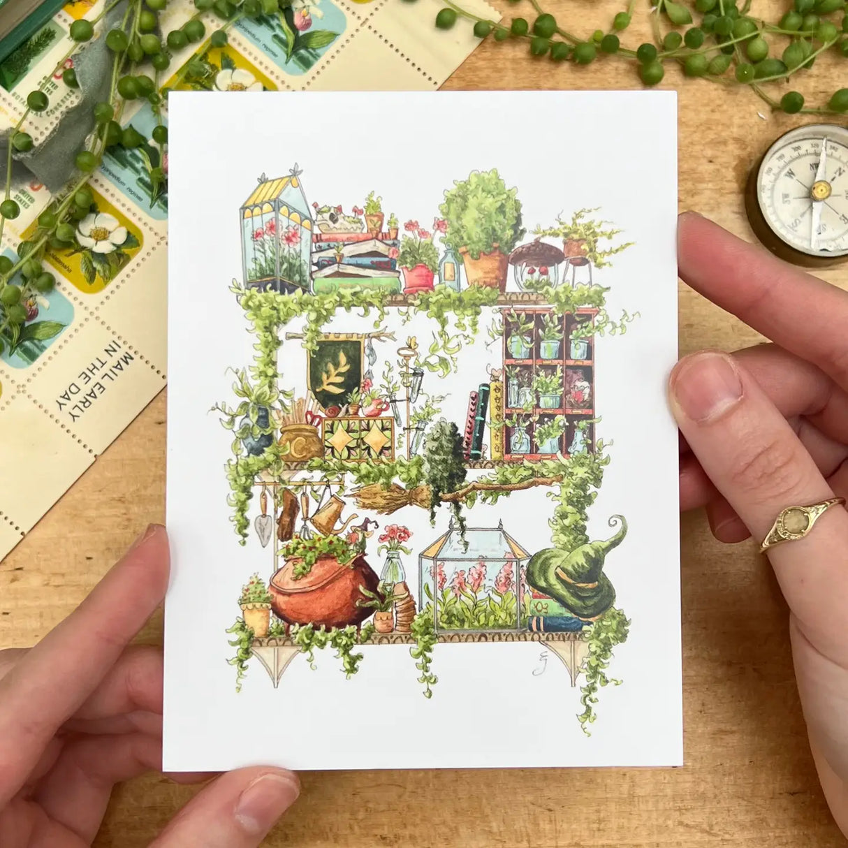 Postcard featuring my original watercolor and ink illustration of the various plants, creatures, and beautiful books nestled on the shelf of a garden witch, including a tiny frog and hedgehog enjoying some itty bitty books, stained glass greenhouses, and endless climbing vines.