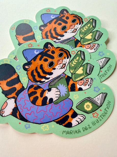 This tiger loves books so much, she can't hold them all at once!

This vinyl matte sticker is weather and waterproof-stick it on a notebook, bike, or car- it's going to stick around! Heck, throw it on a water bottle and through the dishwasher. This sticker can go anywhere. Sticker measures 4" x 4" Printed in USA.