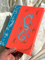 This is a blank 200 page notebook/sketchbook with thin ivory Munken paper perfect for light sketching or writing. Part of a fantasy collection called Woodfolk.

The cover is fluorescent pink/orange and has a smooth matte finish with gold foiling on the front and back. Plus it has a blue fabric spine with white embossing, adding to the charm of the book. Best for pencils, light pens, and generally light mediums, as thick markers or water can bleed. Not recommended for gouache or heavy marker use.