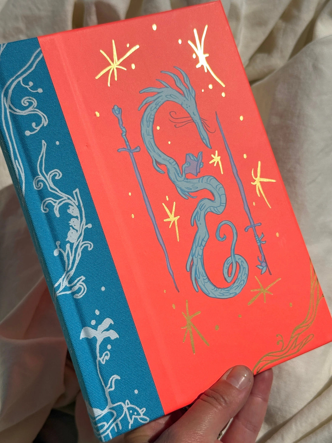 This is a blank 200 page notebook/sketchbook with thin ivory Munken paper perfect for light sketching or writing. Part of a fantasy collection called Woodfolk.

The cover is fluorescent pink/orange and has a smooth matte finish with gold foiling on the front and back. Plus it has a blue fabric spine with white embossing, adding to the charm of the book. Best for pencils, light pens, and generally light mediums, as thick markers or water can bleed. Not recommended for gouache or heavy marker use.