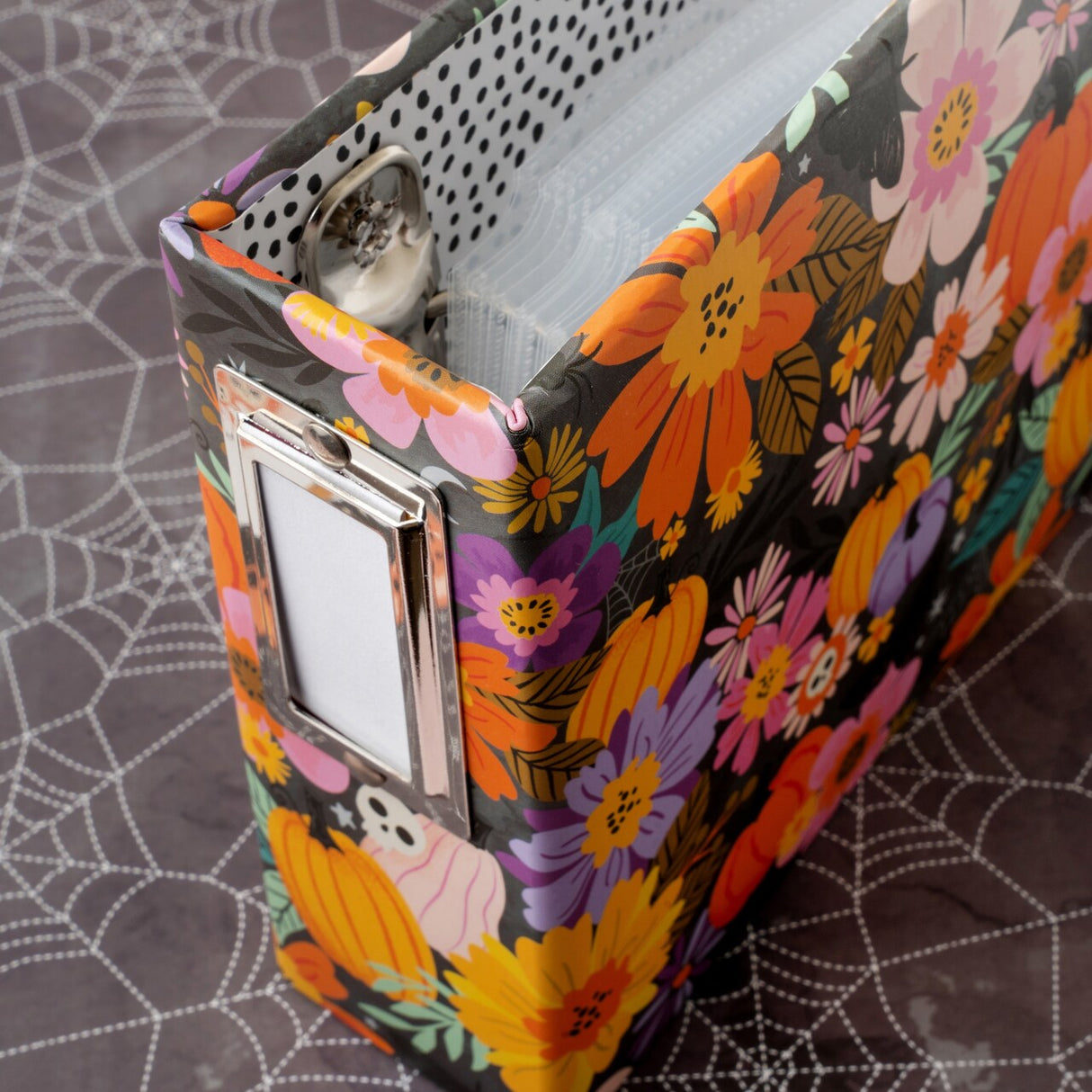 Introducing the 4" x 4" Album from the Tricks And Treats Collection by Paige Evans for American Crafts. This adorable album is the perfect way to showcase your Halloween memories. With its vibrant orange, pink, and purple pumpkins, flowers, skulls, and more on the cover, it captures the festive spirit of the season. The album includes 15 page protectors to keep your precious photos safe and organized. Whether you're documenting a spooky costume party or a fun-filled night of trick-or-treating, this album is