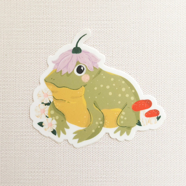 A happy little toad pauses among blooming flowers and mushrooms, a small flower worn proudly atop his head. This vinyl sticker captures that lighthearted moment, celebrating comfort, confidence, and being yourself in the everyday.

Made from durable, waterproof vinyl, this sticker is designed to hold up wherever you place it—whether that’s a water bottle, laptop, or journal you reach for daily.