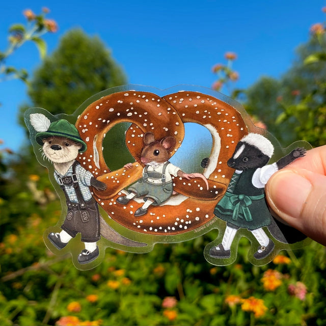 These critters are ready for Oktoberfest! Dressed in their finest lederhosen and dirndl, this otter and skunk are proudly carrying a larger-than-life pretzel. Their mouse pal is just along for the ride!

This die-cut gloss vinyl sticker is durable, weatherproof, and perfect for water bottles, notebooks, laptops, bikes, and all sorts of other surfaces! Make sure to clean and dry the surface thoroughly before applying.