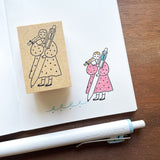 Stamp Marche Maru-Chan Uni-ball Wooden Stamp Rubber stamp with a design of a person holding a thermometer, next to a pink illustration on paper, with a pen on a wooden surface.