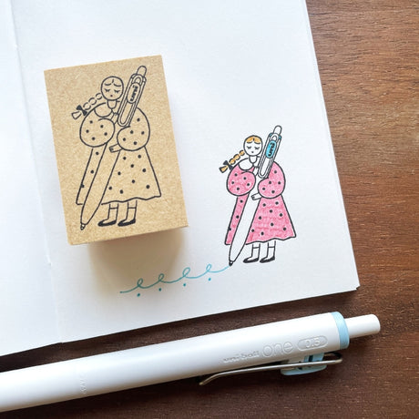 Stamp Marche Maru-Chan Uni-ball Wooden Stamp Rubber stamp with a design of a person holding a thermometer, next to a pink illustration on paper, with a pen on a wooden surface.