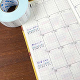 Stamp Marche Planner Label Sticker Roll - Weekly Habit Tracker Calendar with handwritten notes on a wooden surface next to a roll of tape.