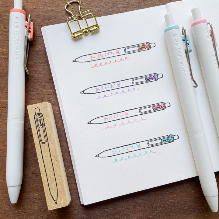 Stamp Marche Uni-ball One Wooden Stamp Notebook with pen illustrations, pens, and a rubber stamp on a wooden surface